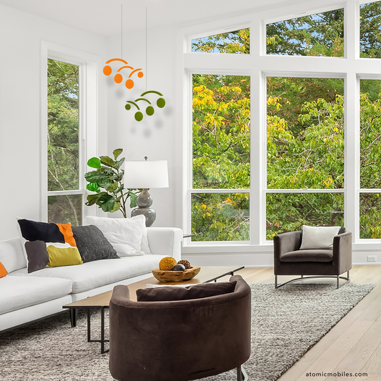 Modern living room with large windows, white walls, and greenery outside and 2 Modernist kinetic hanging art mobiles in orange and olive green in the corner of the room by AtomicMobiles.com