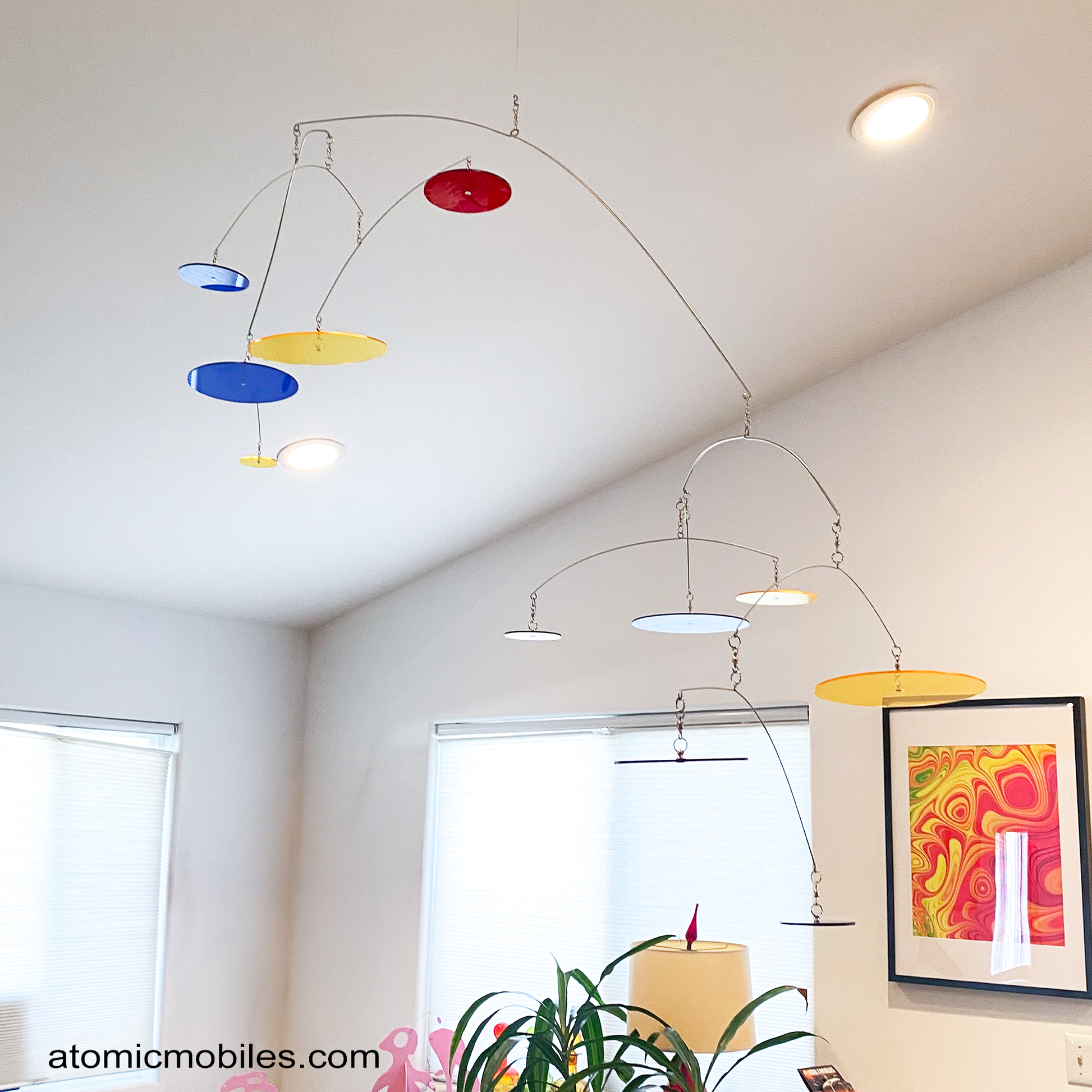Another view of LOOK UP! Art Mobile hanging in colorful living room with abstract art, plant, and windows - mobile with transparent navy, red, and yellow acrylic plexiglass circles by AtomicMobiles.com