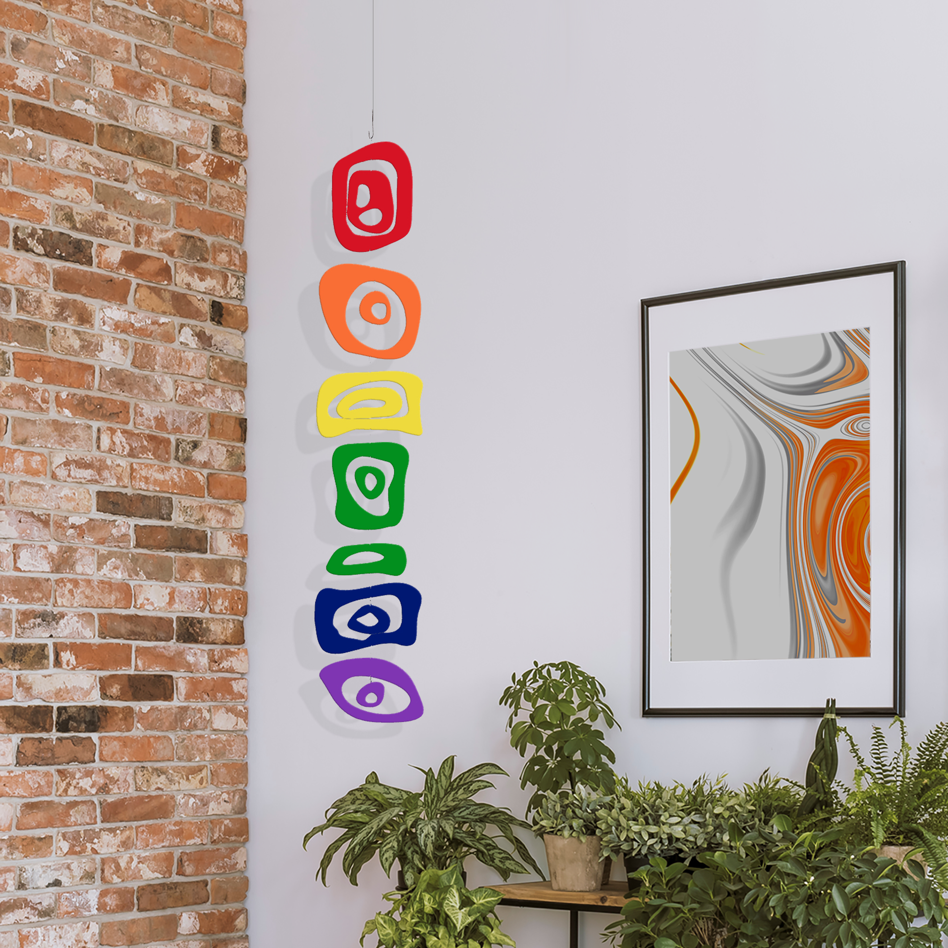 LGBTQ Rainbow Hanging Art Mobile in room with brick wall, plants, and abstract art by AtomicMobiles.com