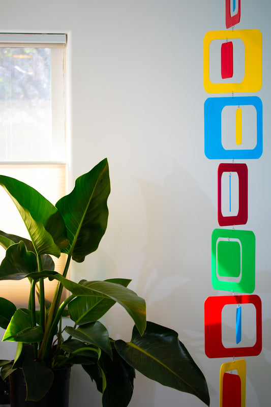 Colorful Vertical Mobile in red, yellow, green, and blue, with large plant next to window - atomicmobiles.com