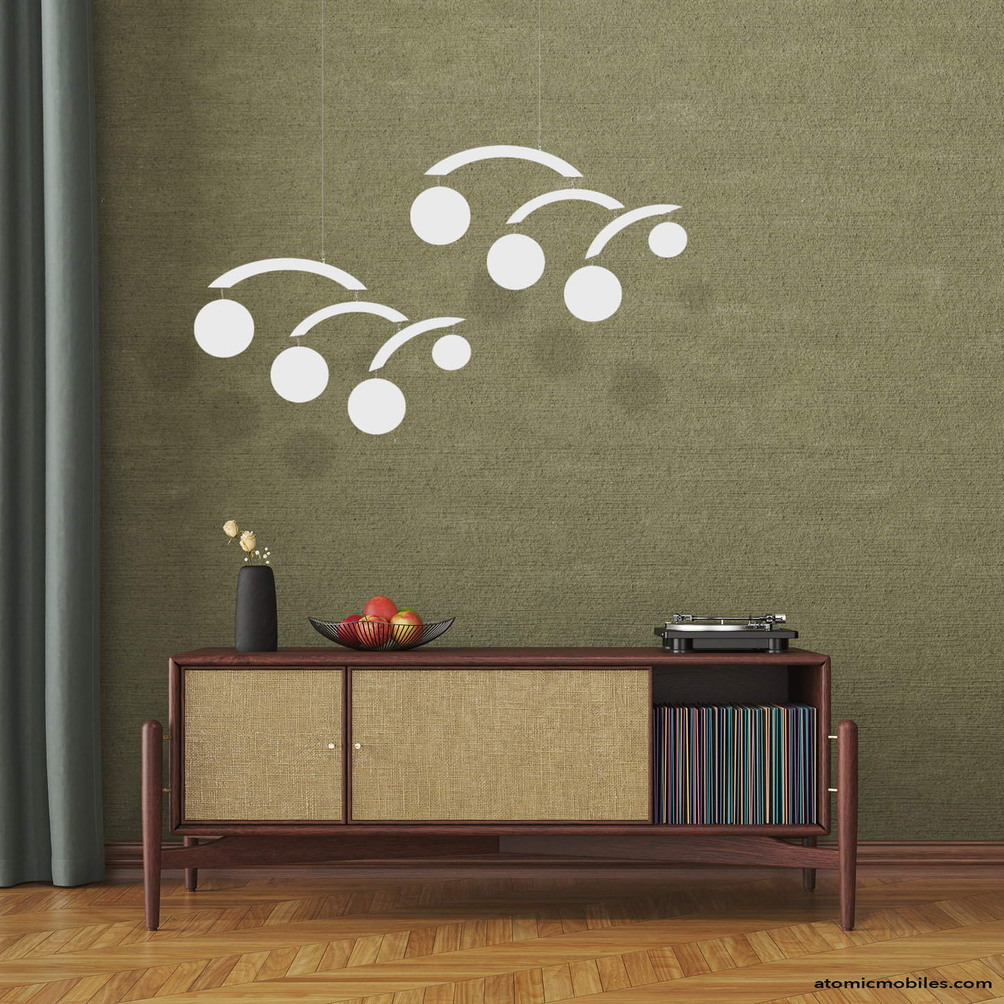 Decorative white mid century modernist hanging art mobiles with circles by AtomicMobiles.com on a green wall above a wooden console table with turntable and vinyl record albums, fruit basket and plant, with parquet floor.