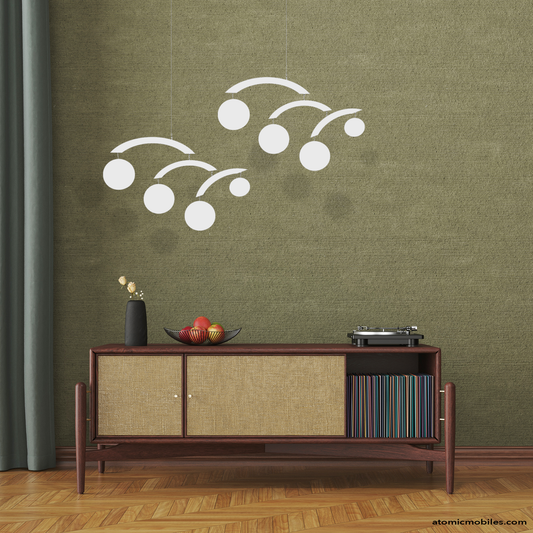 Decorative white mid century modernist hanging art mobiles with circles by AtomicMobiles.com on a green wall above a wooden console table with turntable and vinyl record albums, fruit basket and plant, with parquet floor.