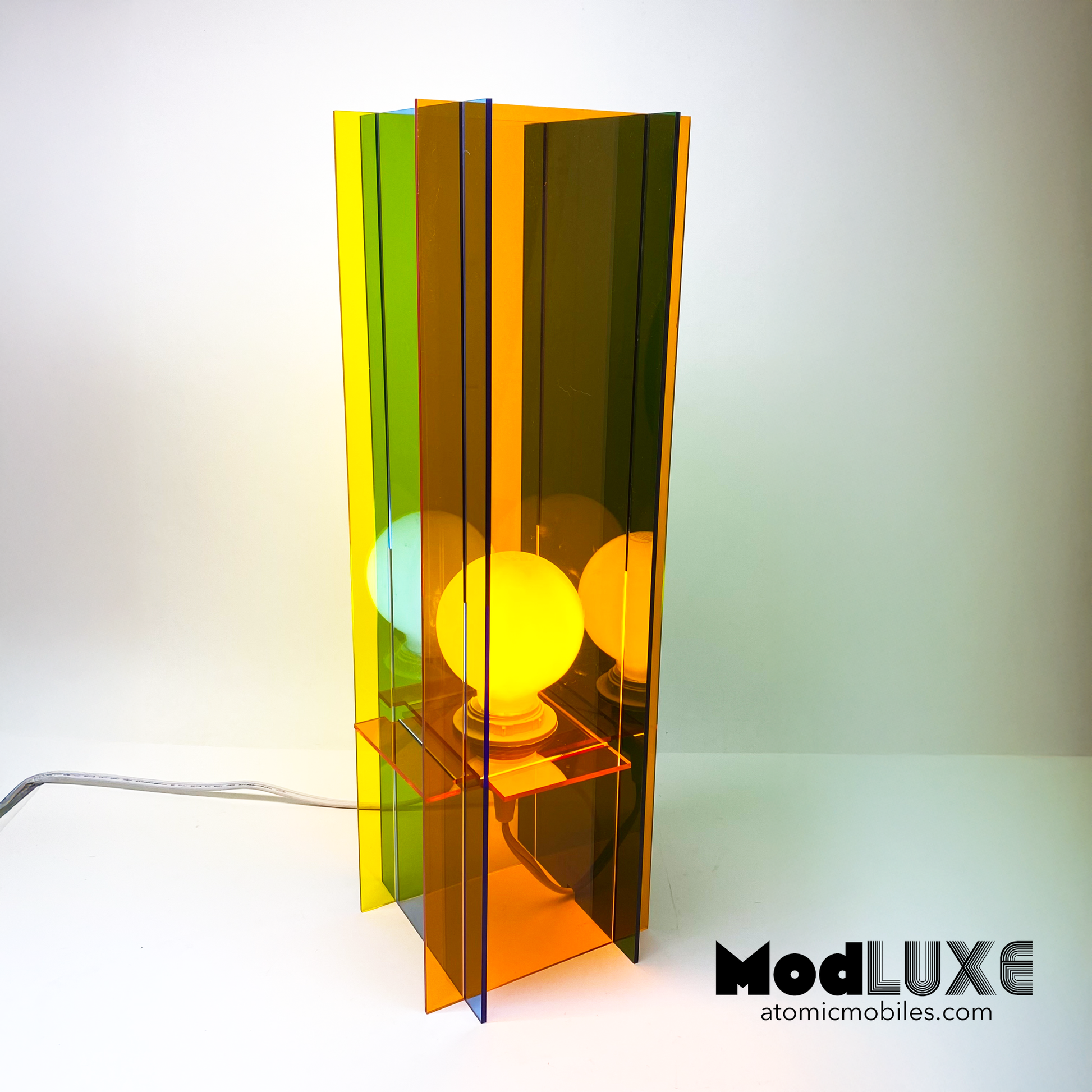 ModLUXE Space Age Lamp in groovy 1970s style and plexiglass acrylic colors of transparent orange, blue, green, and yellow. Colorful lamp with a transparent design on a white background, featuring the brand 'ModLUXE' by AtomicMobiles.com.