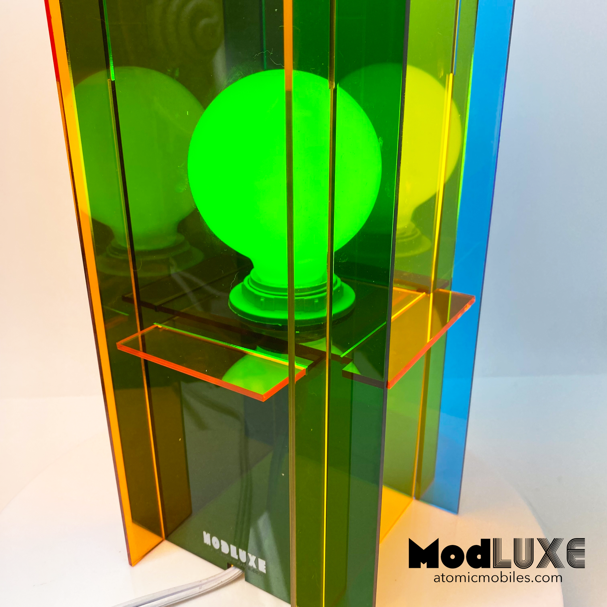 Closeup of ModLUXE Space Age Lamp in groovy 1970s style and plexiglass acrylic colors of transparent orange, blue, green, and yellow. Colorful lamp with a transparent design on a white background, featuring the brand 'ModLUXE' by AtomicMobiles.com.