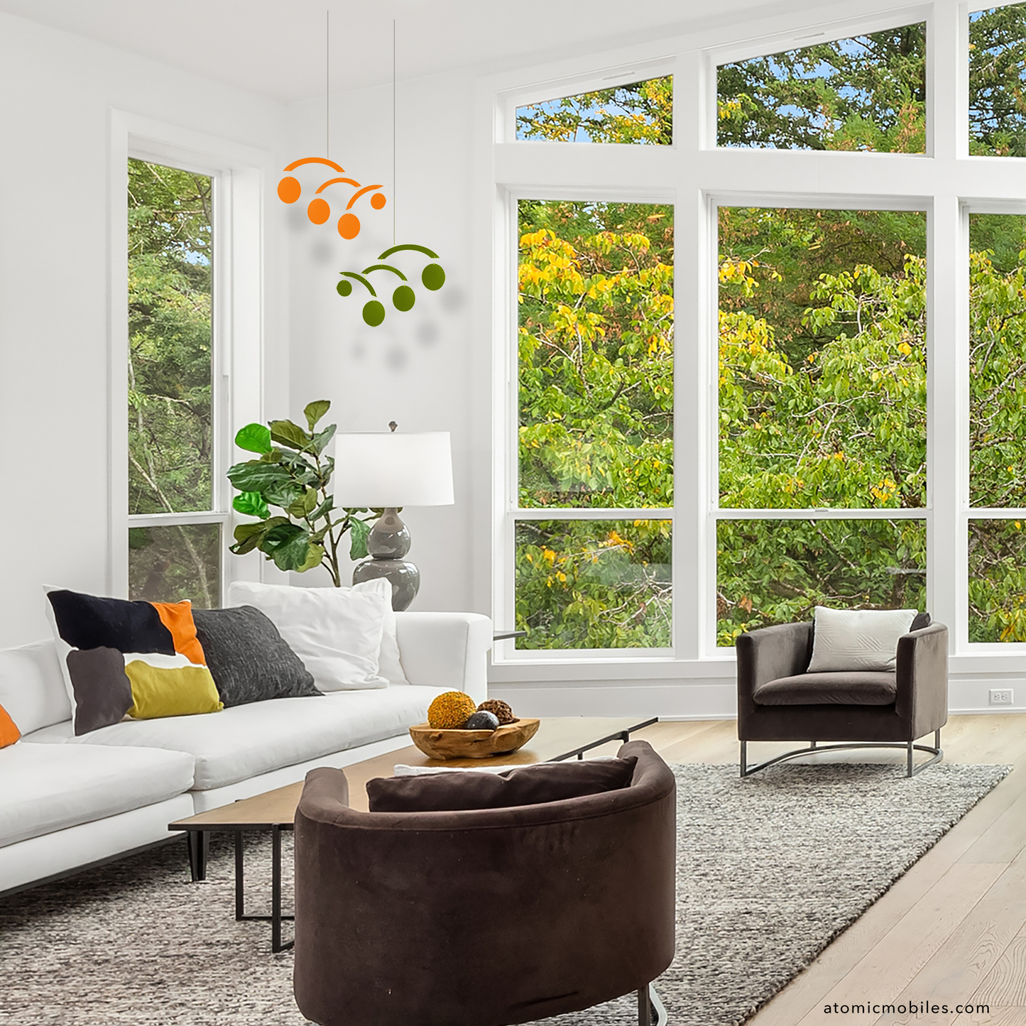 Modern living room with large windows, white walls, and greenery outside and 2 Modernist kinetic hanging art mobiles in orange and olive green in the corner of the room by AtomicMobiles.com
